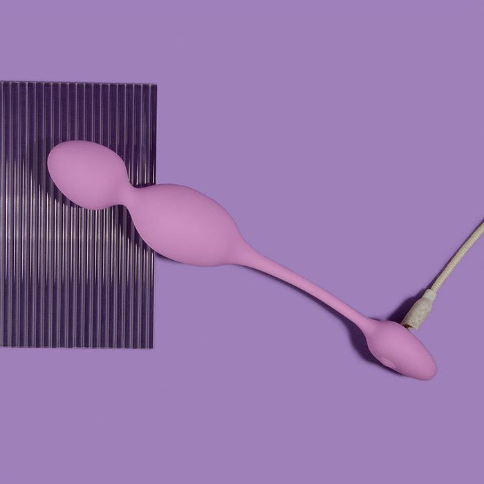 The Raine Vibrating Kegel Ball with Remote Control, a purple silicone object with two bulbous ends and a cord, is placed diagonally on a purple surface. A dark purple textured rectangular sheet is partially visible underneath.