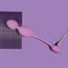 The Raine Vibrating Kegel Ball with Remote Control, a purple silicone object with two bulbous ends and a cord, is placed diagonally on a purple surface. A dark purple textured rectangular sheet is partially visible underneath.