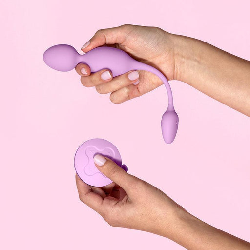 A close-up shows hands holding the Raine Vibrating Kegel Ball with a wireless remote control. This purple pelvic floor strengthener features a smooth finish and is set against a light pink background.
