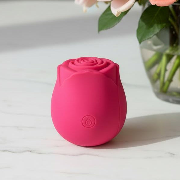 The Ravishing Suction Rose Clitoral Air Pulsation Vibrator, shaped like a pink rose, rests on a white marble surface in front of a glass vase filled with pink and peach roses.