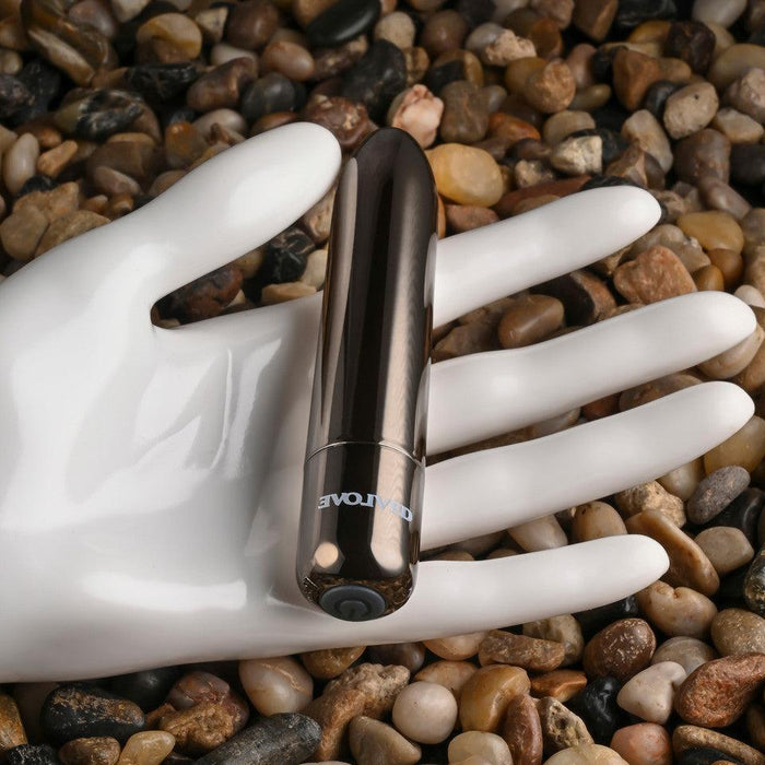 The Real Simple Rechargeable Waterproof Bullet Vibrator in Black Chrome rests on a white mannequin hand atop a surface of smooth, multicolored pebbles.
