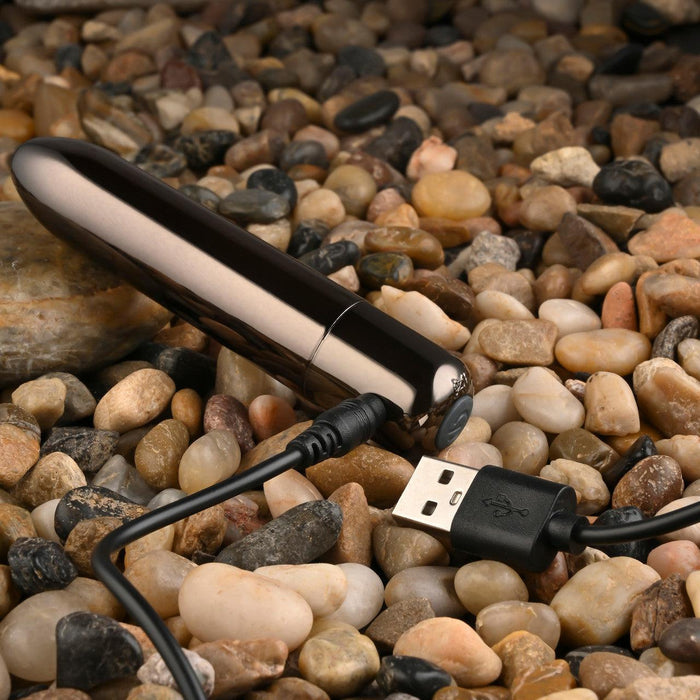The Real Simple Rechargeable Waterproof Bullet Vibrator in Black Chrome, with its USB charging cable, rests on a surface of smooth, multicolored pebbles.