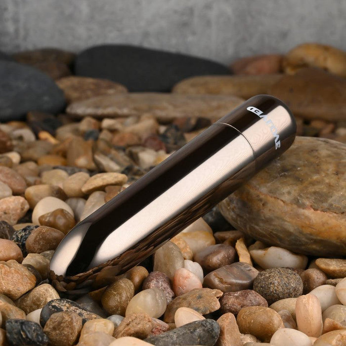 The Real Simple Rechargeable Waterproof Bullet Vibrator in Black Chrome rests on a bed of smooth, multicolored pebbles and stones, showcasing its sleek and luxurious metallic design.