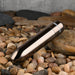 The Real Simple Rechargeable Waterproof Bullet Vibrator in Black Chrome rests on a bed of smooth, multicolored pebbles and stones, showcasing its sleek and luxurious metallic design.