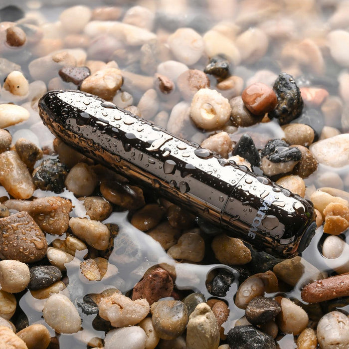 The Real Simple Rechargeable Waterproof Bullet Vibrator in Black Chrome rests among wet pebbles and rocks, its shiny, black cylindrical surface glistening with water drops as it lies partially submerged in shallow water.