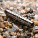 The Real Simple Rechargeable Waterproof Bullet Vibrator in Black Chrome rests among wet pebbles and rocks, its shiny, black cylindrical surface glistening with water drops as it lies partially submerged in shallow water.