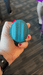 A hand with red-painted nails holds the Riana Circular Clitoral Rubbing Vibrator, made of body-safe teal silicone. The person wears a black watch, and the carpeted floor and parts of people's legs are visible in the background.