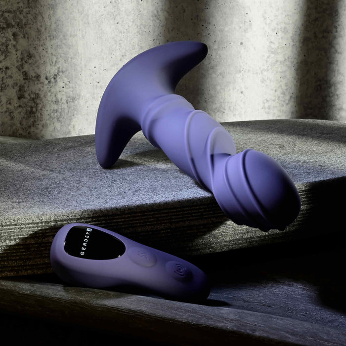 The Ring It Remote Control Rimming Vibrating Prostate Plug, a purple massager with a curved base and textured shaft, rests on a gray stone surface next to its matching remote control.