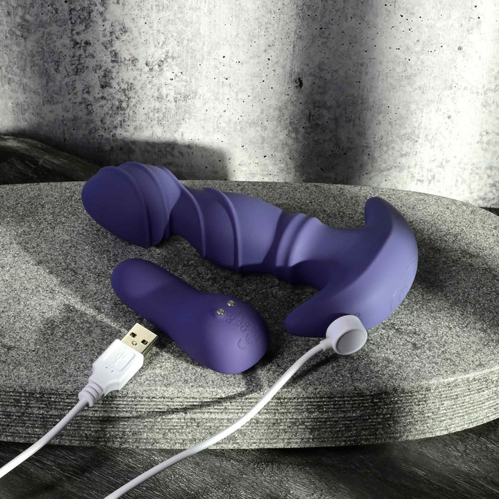 The Ring It Remote Control Rimming Vibrating Prostate Plug, made of purple silicone with a curved, ribbed design, rests on a gray stone surface alongside its remote control and USB charging cable.
