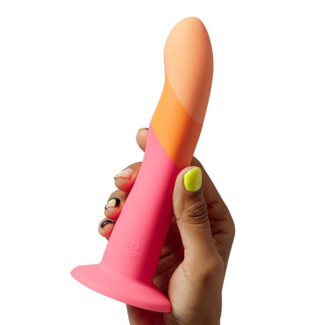 A hand with brightly painted nails holds the Romp Dizi Ultra Smooth 7 Inch G-Spot Dildo With Suction Cup, a colorful silicone toy with pink, orange, and peach tones, designed for p-spot and g-spot stimulation and fully harness compatible.