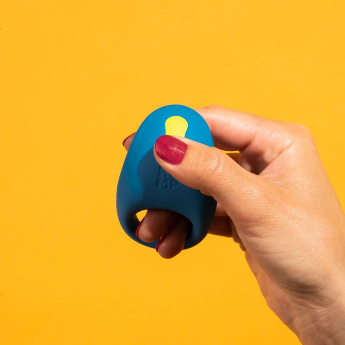 A hand with red-painted nails holds the Romp Juke Beginner-Friendly Vibrating Couples Cock Ring with a yellow button against a bright yellow background, ideal for enhancing couples’ intimacy.