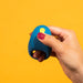A hand with red-painted nails holds the Romp Juke Beginner-Friendly Vibrating Couples Cock Ring with a yellow button against a bright yellow background, ideal for enhancing couples’ intimacy.
