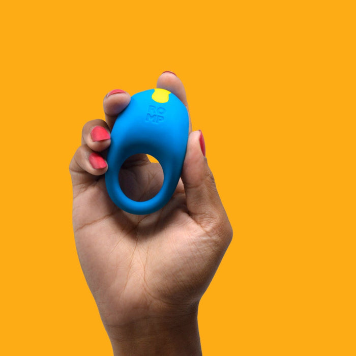 A hand with red-painted nails holds the Romp Juke Beginner-Friendly Vibrating Couples Cock Ring, featuring a blue design and yellow button, against an orange background to enhance intimacy and support erection firmness.