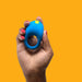 A hand with red-painted nails holds the Romp Juke Beginner-Friendly Vibrating Couples Cock Ring, featuring a blue design and yellow button, against an orange background to enhance intimacy and support erection firmness.