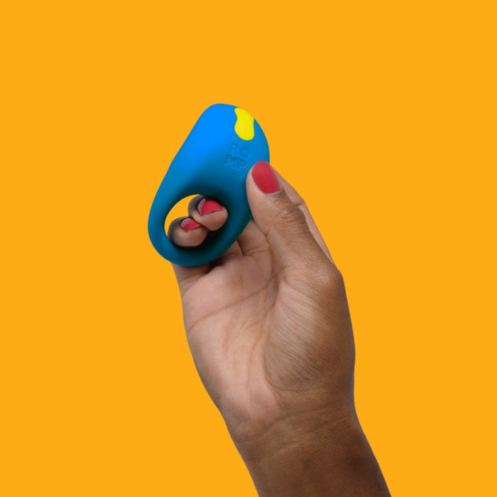 A hand with red-painted nails holds the Romp Juke Beginner-Friendly Vibrating Couples Cock Ring, blue and yellow in color, embossed with "ROMP," against an orange background—designed to enhance intimacy and playful excitement.