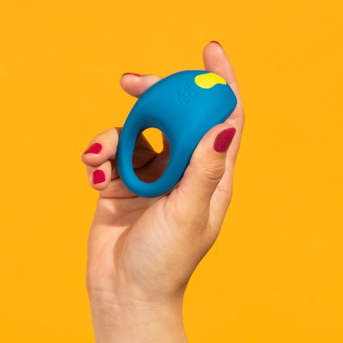 A hand with red nail polish holds the Romp Juke Beginner-Friendly Vibrating Couples Cock Ring, featuring a blue design with a yellow accent, against a solid orange background.