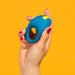 A hand with red nail polish holds the Romp Juke Beginner-Friendly Vibrating Couples Cock Ring, featuring a blue design with a yellow accent, against a solid orange background.