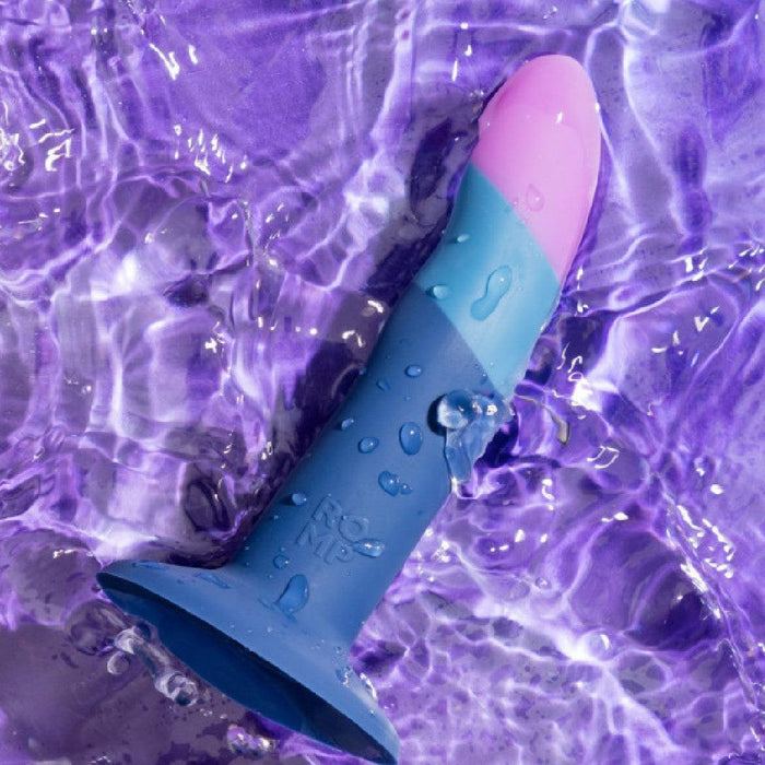 The Romp Piccolo Sleek First Time 5 Inch Silicone Dildo with Suction Cup, in blue and pink, rests on a purple water-textured surface, glistening with droplets—ideal for anal play or versatile G-spot stimulation.