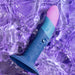The Romp Piccolo Sleek First Time 5 Inch Silicone Dildo with Suction Cup, in blue and pink, rests on a purple water-textured surface, glistening with droplets—ideal for anal play or versatile G-spot stimulation.