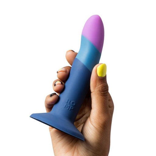 A hand with painted nails holds the Romp Piccolo Sleek First Time 5 Inch Silicone Dildo with Suction Cup, a colorful blue, purple, and pink silicone toy featuring a flared base and "ROMP" branding—ideal for anal play.