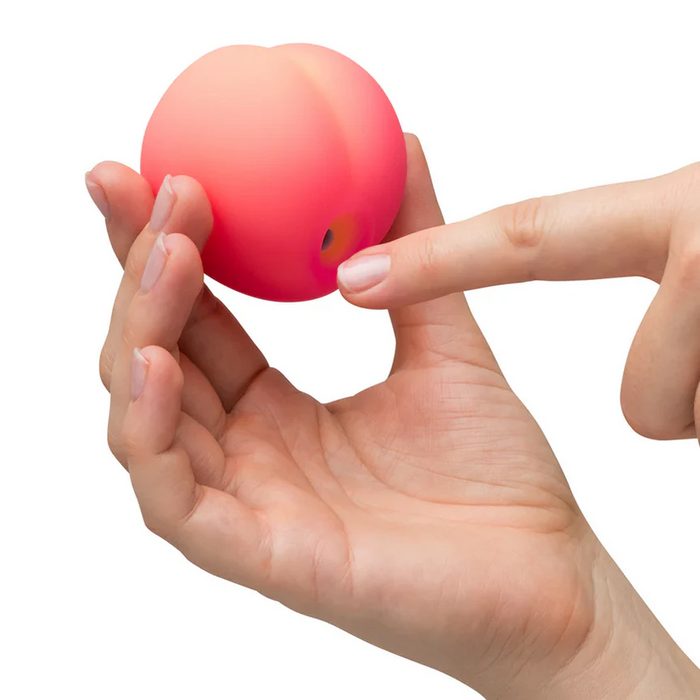 A hand holds the Romp Pink Peach Pleasure Air Clitoris Stimulator, a small, peach-shaped toy with a hole at the bottom. Another hand's finger touches the hole. The plain white background highlights this beginner-friendly device.