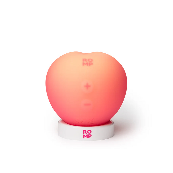 The Romp Pink Peach Pleasure Air Clitoris Stimulator—a smooth, pink, peach-shaped toy with control symbols—stands on a white ROMP base and offers beginner-friendly, playful air pleasure for exploring new sensations.