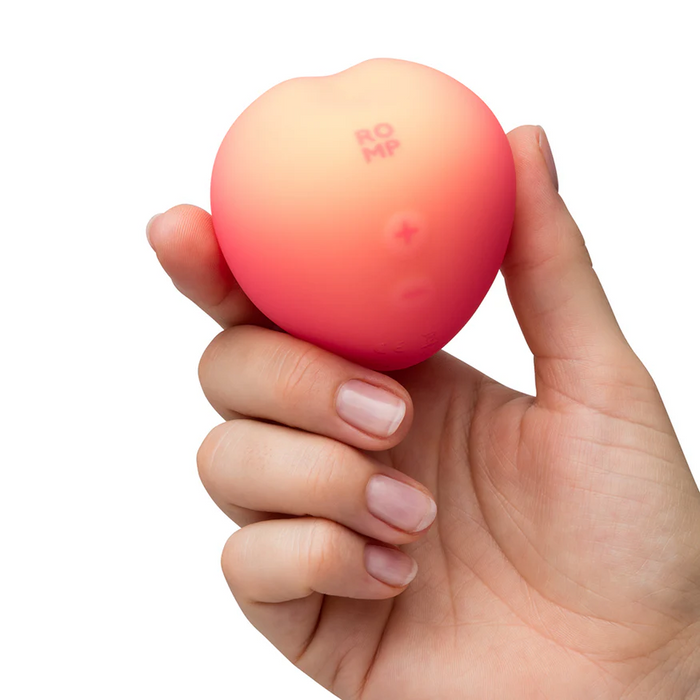 A hand holds the Romp Pink Peach Pleasure Air Clitoris Stimulator, a small, round, peach-colored device with "ROMP" and two raised symbols on its smooth surface, designed for beginners and shown against a white background.