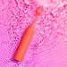 The Romp Pop Pinpoint Clitoral Stimulator with 2 Heads, in coral, features water droplets on its surface as it rests on rippling water reflecting pink light.