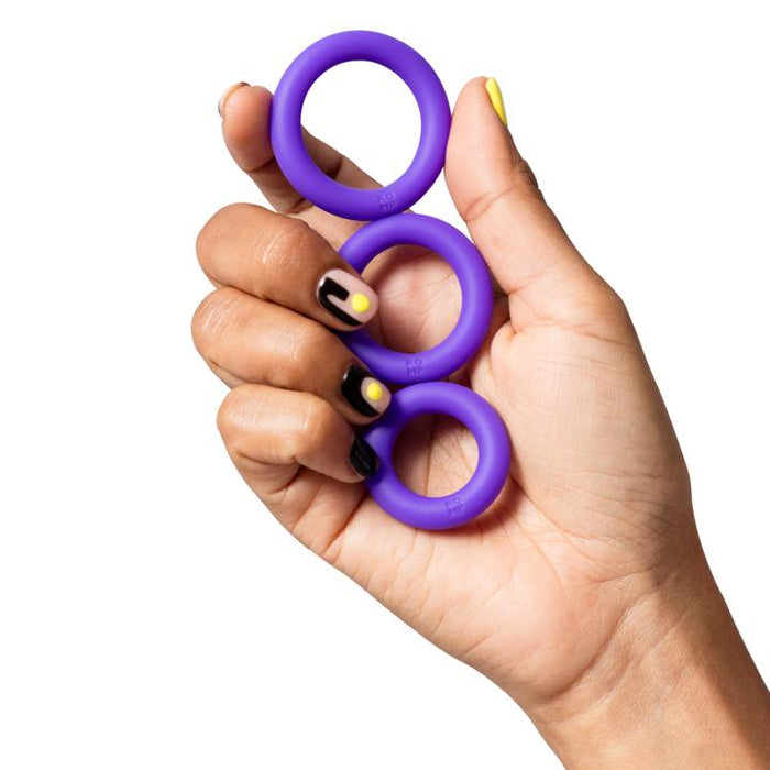 A hand with black and yellow painted nails holds the Romp Remix Easy to Use Cock Ring Trio—three stretchy silicone rings of different sizes—against a white background.