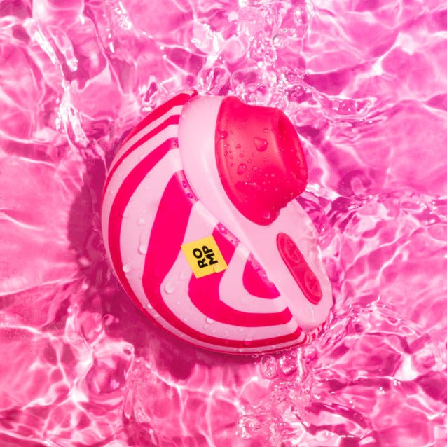 The Romp Spin Small Discreet Pleasure Air Clitoral Stimulator, with pink and white stripes and a yellow label, floats in rippling pink water, casting playful reflections.
