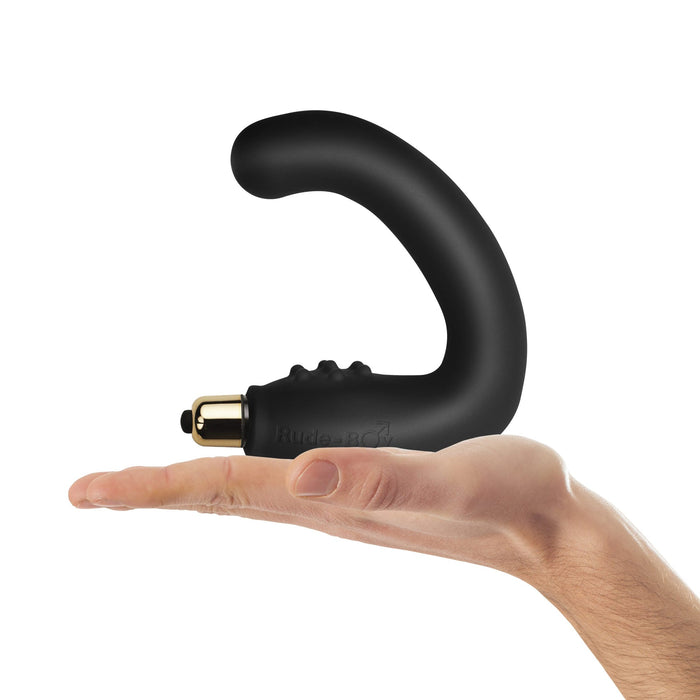 A hand holds the Rude-Boy Dual Prostate & Perineum Stimulator, a black massager with a gold-colored tip, against a plain white background.