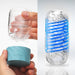 Two hands hold parts of the Tenga Spinner DX Drops Textured Masturbator—a transparent, blue-based male stroker. Next to it, the toy is fully assembled, highlighting its blue spiral design, textured interior, and thicker elastomer against a white background.