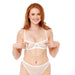 A woman with long red hair smiles in a sheer white bra and matching underwear, subtly showcasing the Peaches 'n Cream Pearl Nipple Clamps, which add a touch of elegance against the plain white background.