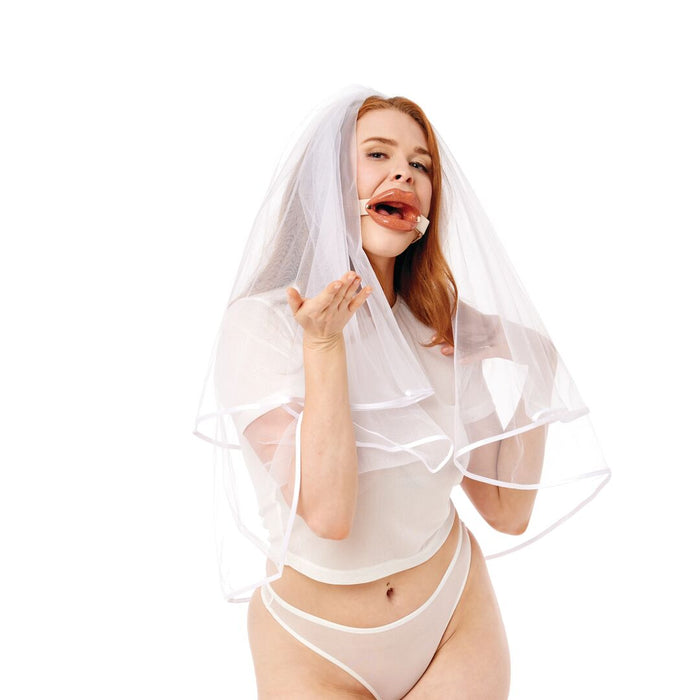 A woman in white lingerie and a bridal veil playfully blows a kiss while modeling the Peaches 'n Cream Silicone Lips Open Mouth Gag against a plain white background.