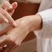 Wearing a white robe, a person applies Sex au Naturel Clitoral Arousal Serum—a clear gel—to the back of their hand with their fingertip, while a softly blurred background creates a soothing atmosphere.