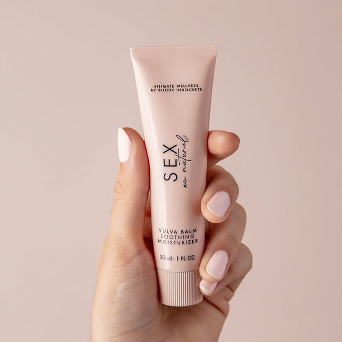 A hand with light pink nails holds a small pink tube labeled "Sex au Naturel Vulva Balm Soothing Moisturizer," the ideal menopause moisturizer for intimate wellness, set against a soft pink background.