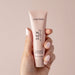 A hand with light pink nails holds a small pink tube labeled "Sex au Naturel Vulva Balm Soothing Moisturizer," the ideal menopause moisturizer for intimate wellness, set against a soft pink background.