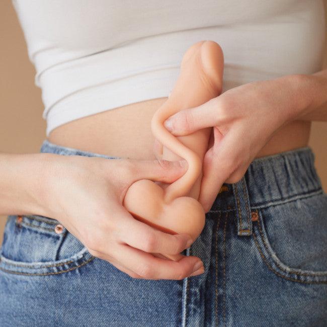 A person in a white crop top and blue jeans holds the Sliding Skin Realistic Small 7 Inch Light Tone Silicone Dildo with Suction Cup at waist level with both hands.