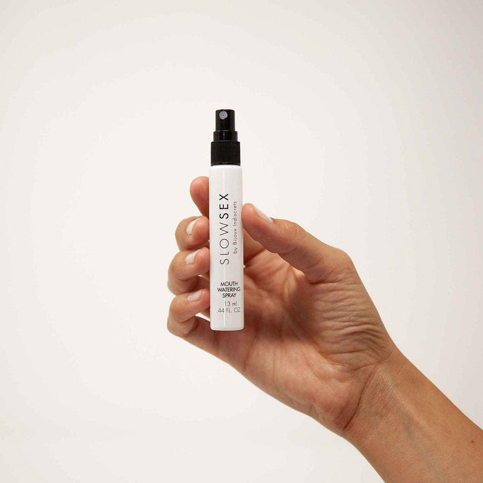 A hand holds a small spray bottle labeled "Slow Sex Mouthwatering Spray" by Bijoux Indiscrets against a plain, light background. The bottle is white with black text and has a black spray nozzle, designed to enhance saliva production during oral sex sessions.