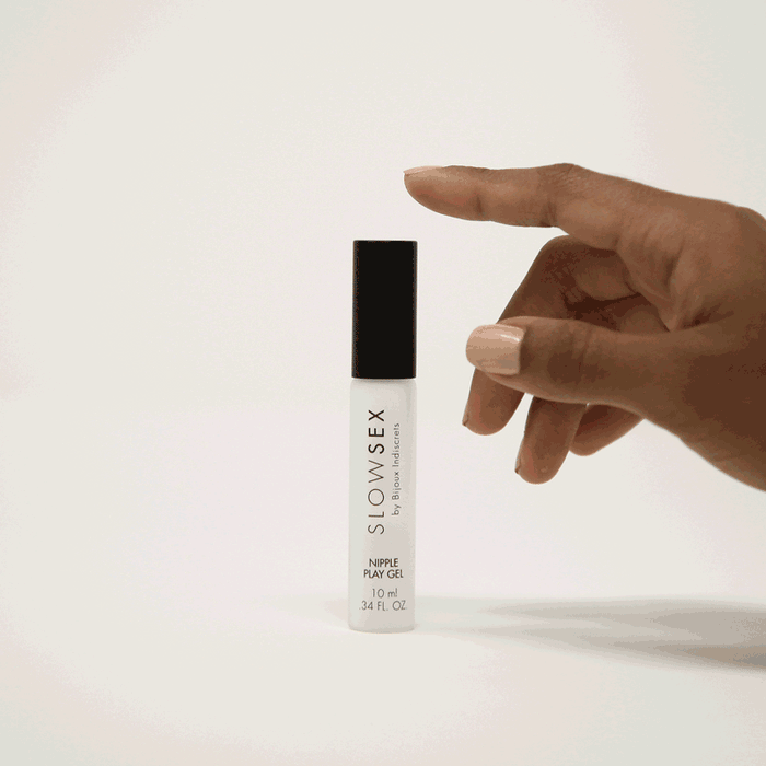 A hand with light-colored nails is about to flick a small, clear bottle of Slow Sex Nipple Play Gel, featuring a black cap and roll-on applicator, against a plain white background.