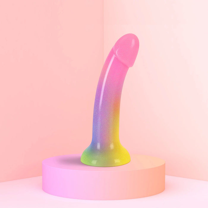 The Stargazer Smooth 7 Inch Rainbow Ombre Glitter Silicone Dildo with Suction Cup shines in pink, purple, blue, and yellow tones as it stands upright on a pink platform against soft pink walls—a perfect choice for pegging or g-spot play.