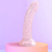 The Starlight Smooth 7 Inch Pink Glitter Silicone Dildo with Suction Cup, featuring a curved shape, stands upright on a pastel pedestal against a soft purple background.