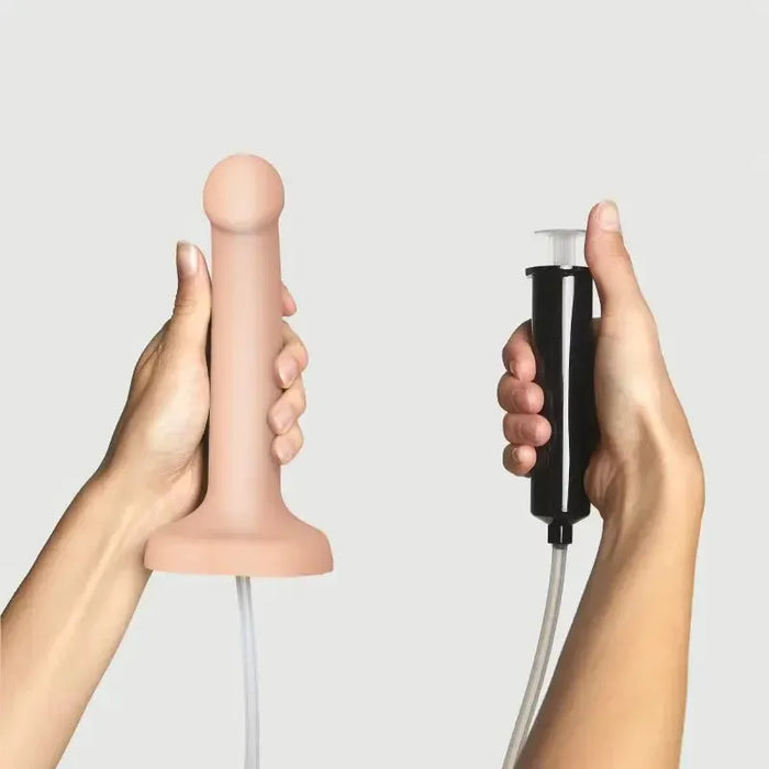 A person holds the Strap-On-Me 7.5 Inch Silicone Squirting Dildo - Vanilla in one hand and a black hand pump with tube in the other, set against a plain light background.