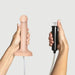 A person holds the Strap-On-Me 7.5 Inch Silicone Squirting Dildo - Vanilla in one hand and a black hand pump with tube in the other, set against a plain light background.