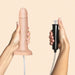 A hand holds the Strap-On-Me Realistic 7.5 Inch Silicone Squirting Dildo - Vanilla with an attached tube, while another hand holds a black handheld pump connected by the tube, against a light peach background.