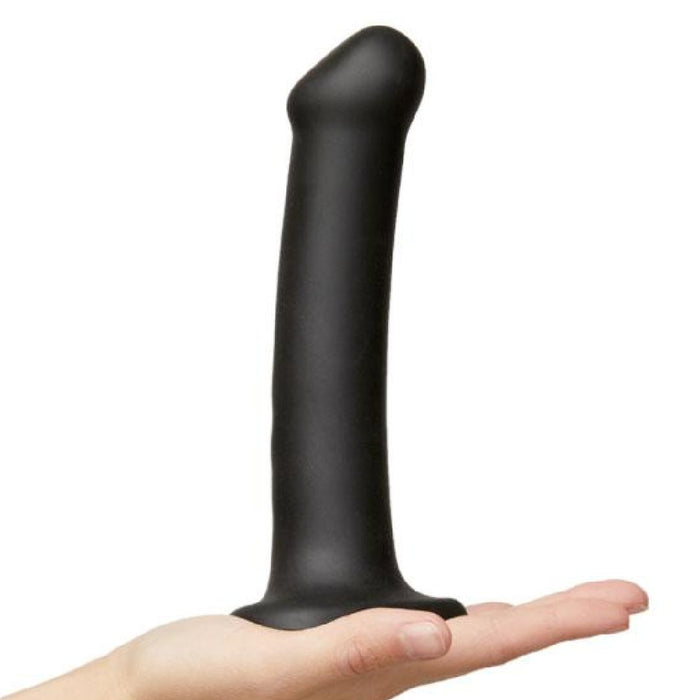 A hand holds the Strap-On-Me Smooth Semi-Realistic 8 Inch Black Silicone Dildo, featuring a large size and flared base for harness compatibility, against a plain white background.