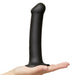 A hand holds the Strap-On-Me Smooth Semi-Realistic 8 Inch Black Silicone Dildo, featuring a large size and flared base for harness compatibility, against a plain white background.