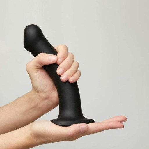 Someone holds the Strap-On-Me Smooth Semi-Realistic 8 Inch Black Silicone Dildo upright, supporting its base against a light background—ideal for use with a strap-on harness or for g-spot and prostate stimulation.