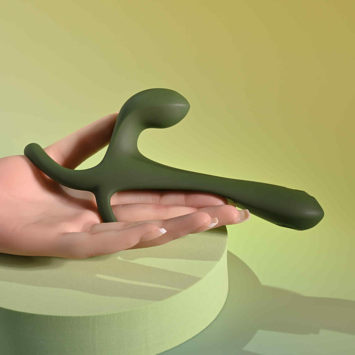 A hand holds the Strap Tap & Go Girth Enhancer & Tapping Vibrator—a sleek, green, modern couples sex toy with smooth curves and matte finish—set against a soft gradient background in green and yellow tones.