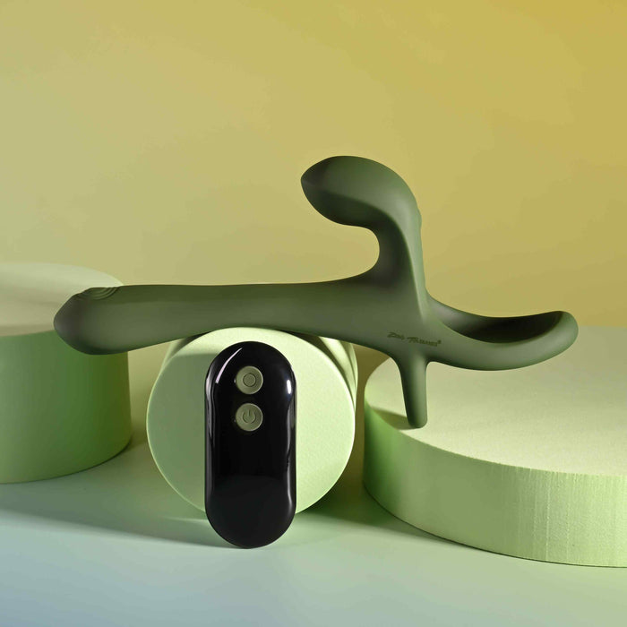 The Strap Tap & Go Girth Enhancer & Tapping Vibrator, a green silicone hands-free couples toy with an ergonomic shape, rests on light green cylinders beside its two-button black remote, against a soft yellow and green background.